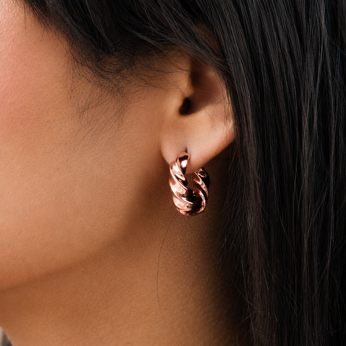 A close-up of a person's ear wearing a twisted hoop earring in a polished rose gold finish, with dark hair framing the side of the face.