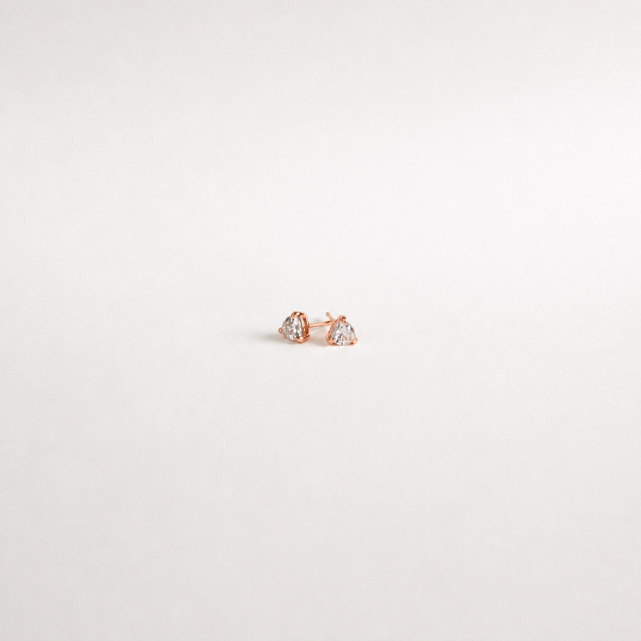 A pair of small rose gold stud earrings with clear heart-shaped gemstones, displayed on a clean, light off-white background.