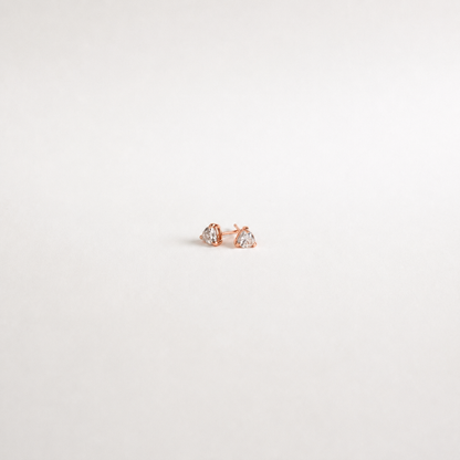 A pair of small rose gold stud earrings with clear heart-shaped gemstones, displayed on a clean, light off-white background.