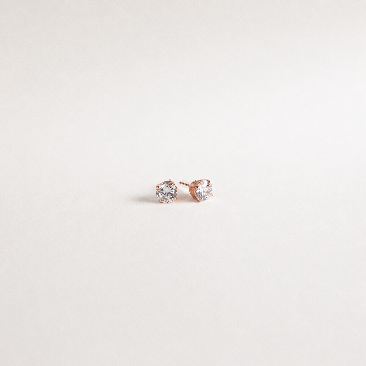 A pair of small round stud earrings in rose gold with clear gemstones, displayed on a clean, light off-white background.