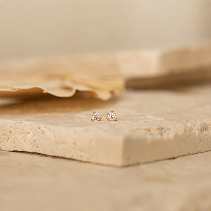 A pair of small gold stud earrings with clear gemstones, resting on a textured beige stone slab with a soft-focus background.