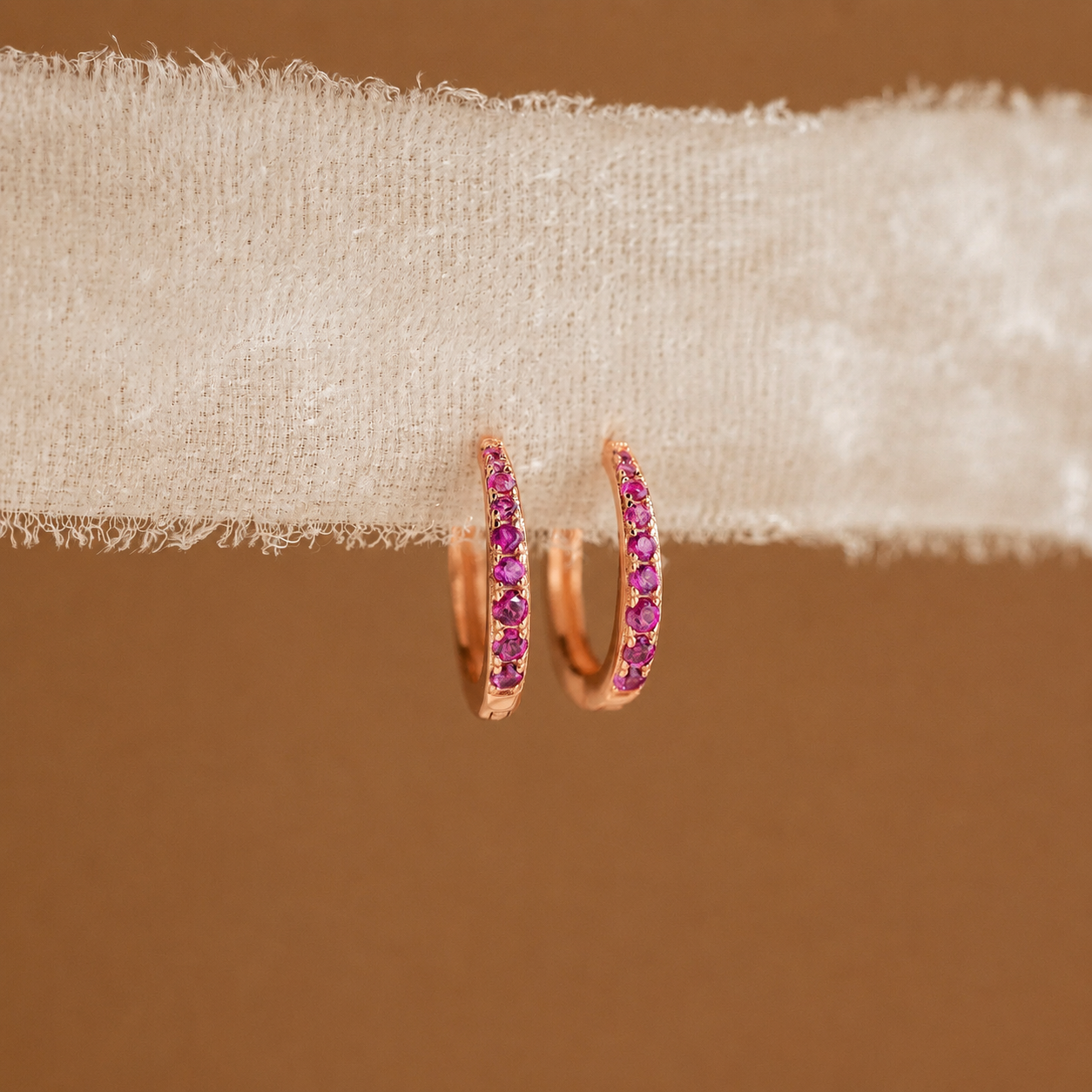 Rose gold hoop earrings with pink gemstones on beige fabric against a brown background