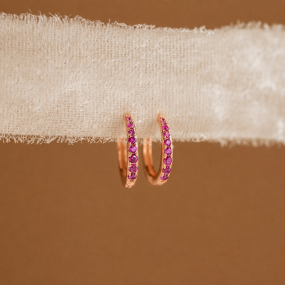 Rose gold hoop earrings with pink gemstones on beige fabric against a brown background