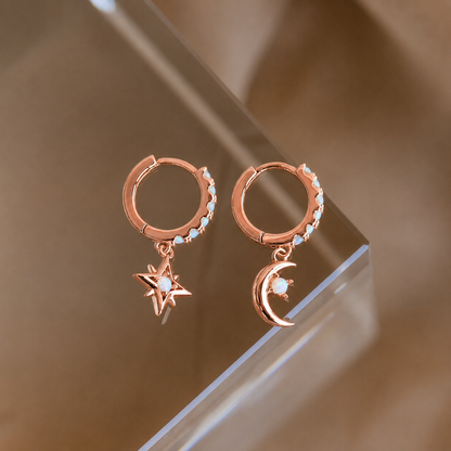 Rose gold hoop earrings with star and moon charms on a reflective surface.