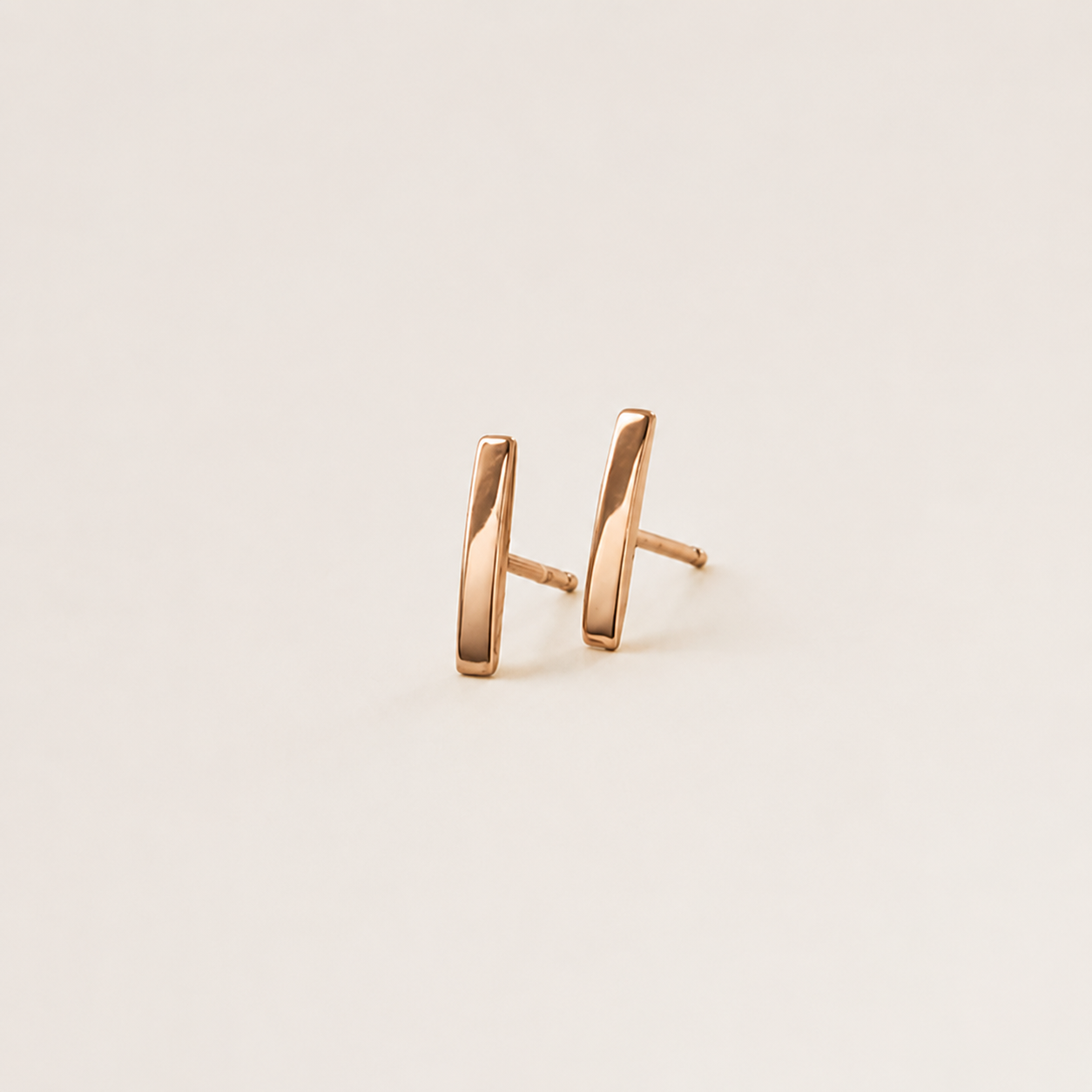 A pair of minimalist rose gold stud earrings with a polished, slightly angled rectangular bar design, shown on a clean off-white background.
