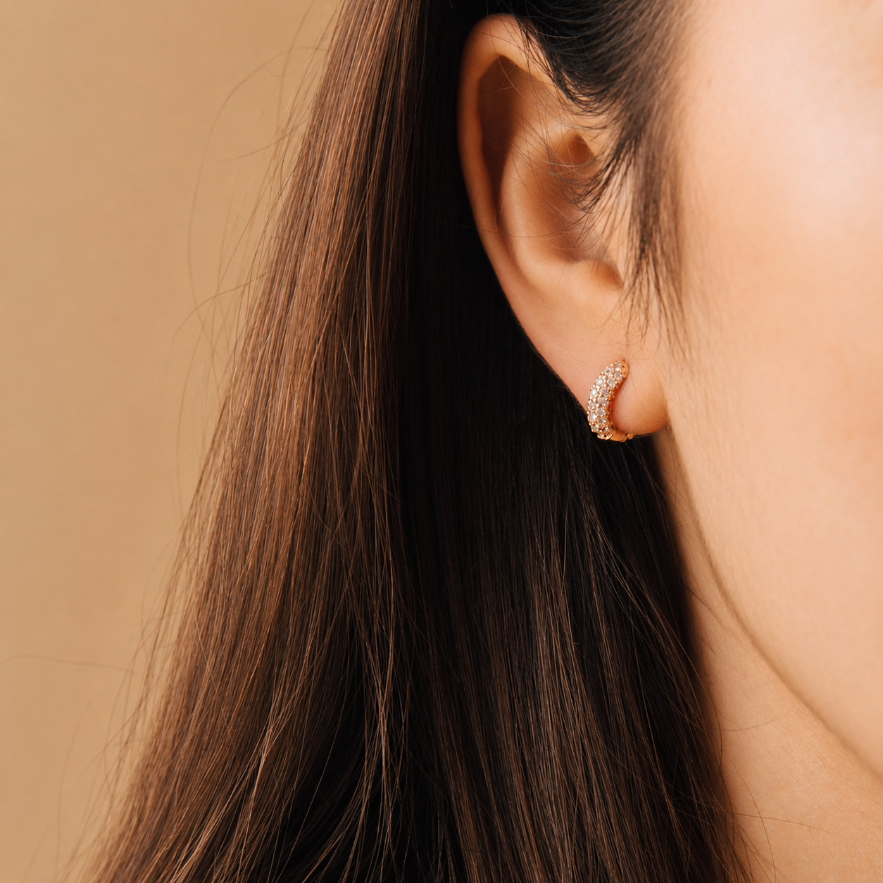 A close-up of a model's ear wearing a small crescent-shaped hoop earring set with clear stones on a warm beige background.