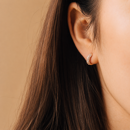 A close-up of a model's ear wearing a small crescent-shaped hoop earring set with clear stones on a warm beige background.