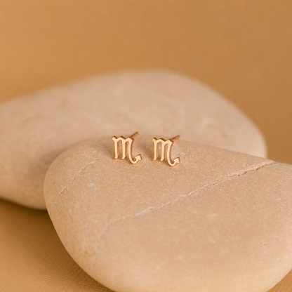 Gold 'm' earrings on a beige stone with a beige background