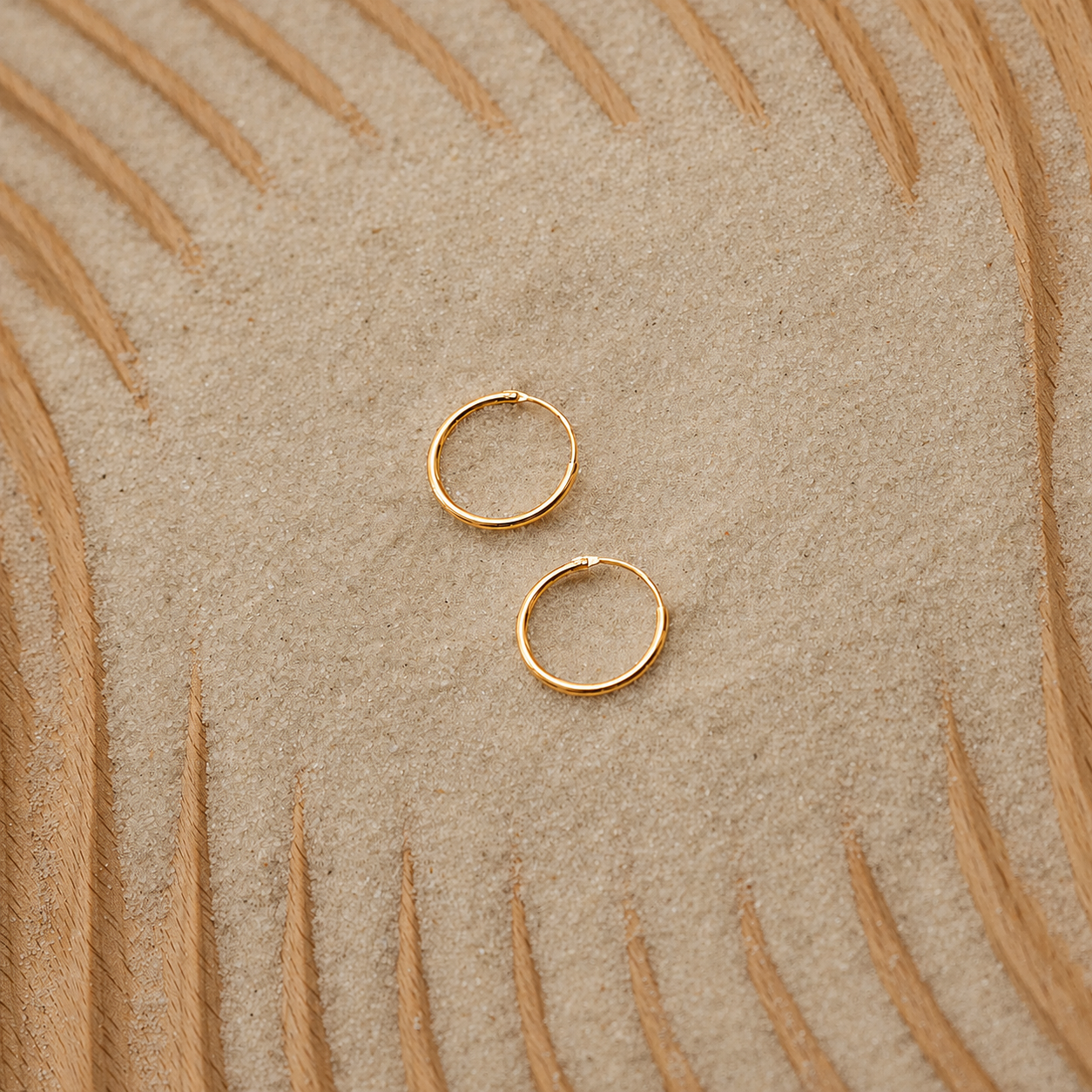 Two small gold hoop earrings placed on a textured beige surface with a curved tan pattern in the background.