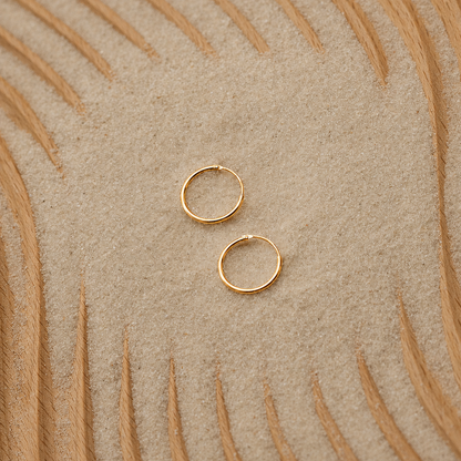 Two small gold hoop earrings placed on a textured beige surface with a curved tan pattern in the background.