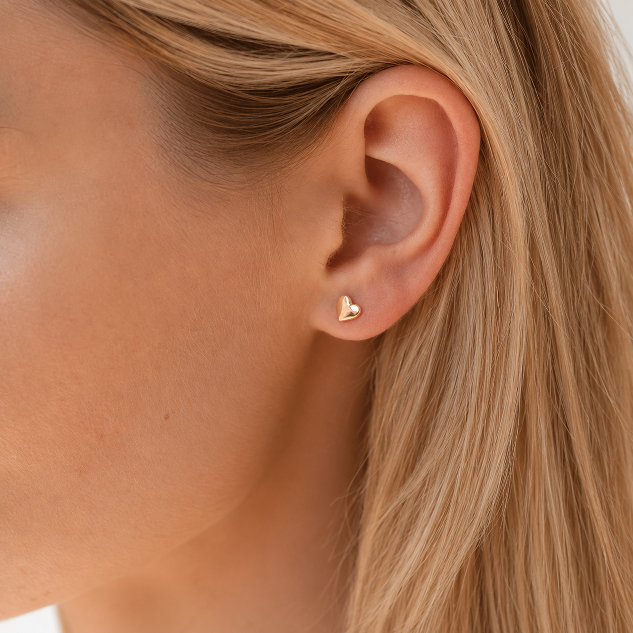 Close-up of an ear wearing a gold heart-shaped earring with blonde hair.