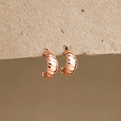 A pair of small curved hoop earrings in a warm metallic pinkish-gold tone, photographed against a split beige and taupe background.