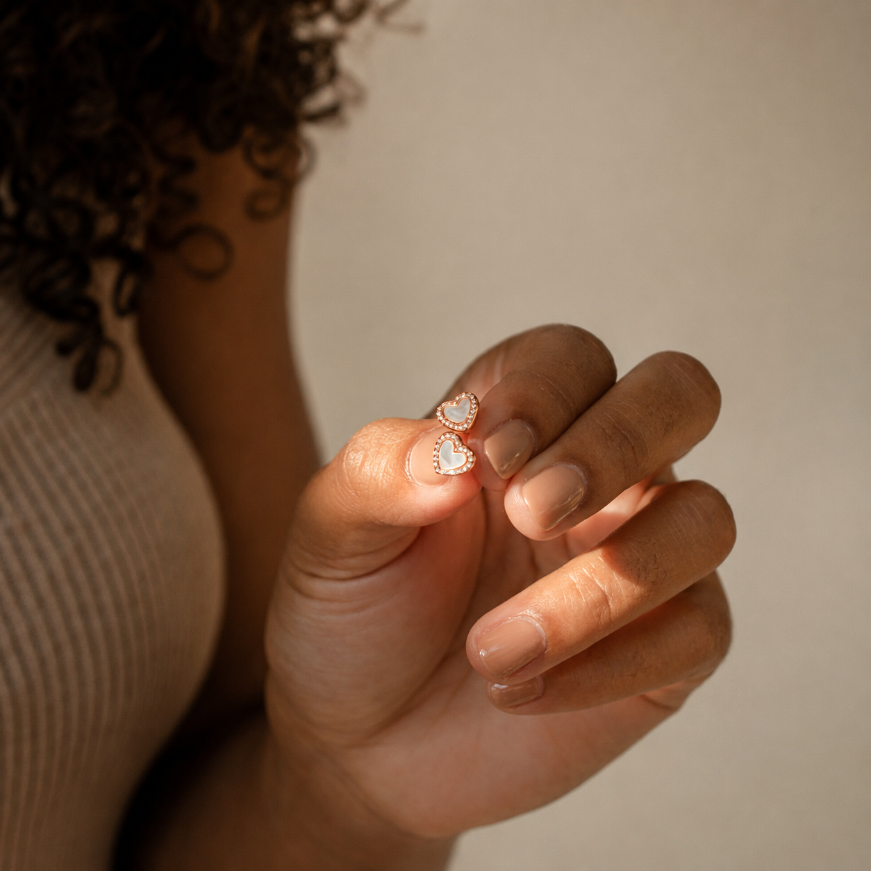 A close-up of a person's hand holding a pair of small heart-shaped stud earrings with white stones and gold settings, against a soft beige background.