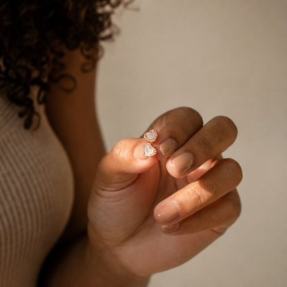 A close-up of a person's hand holding a pair of small heart-shaped stud earrings with white stones and gold settings, against a soft beige background.