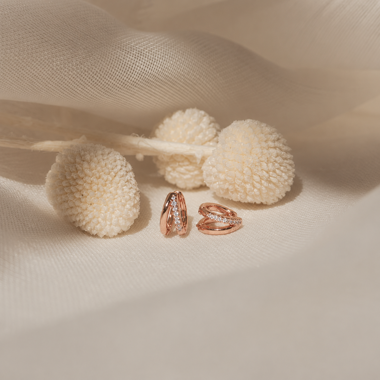 A pair of rose gold hoop earrings with small clear stones, displayed on a soft beige fabric next to dried cream-colored pom-pom flowers.