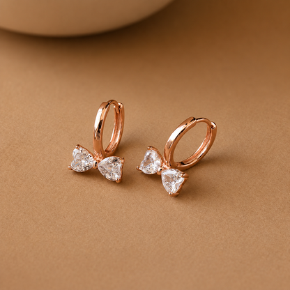 Rose gold bow-shaped earrings with clear gemstones on a brown background