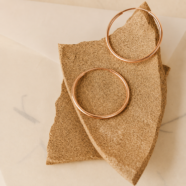 Two thin rose gold hoop earrings displayed on a textured tan stone slab over a light marble surface.