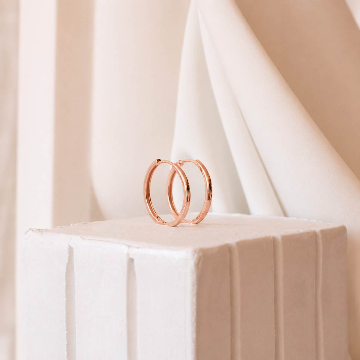 Rose gold hoop earrings on a white pedestal with a soft beige background