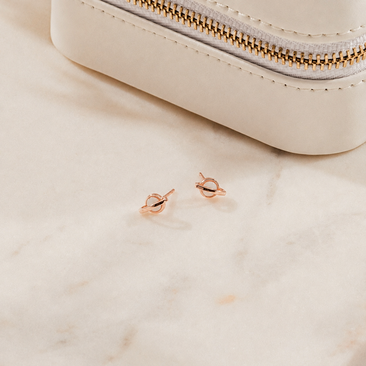 Rose gold earrings on a beige marble surface with a beige pouch.