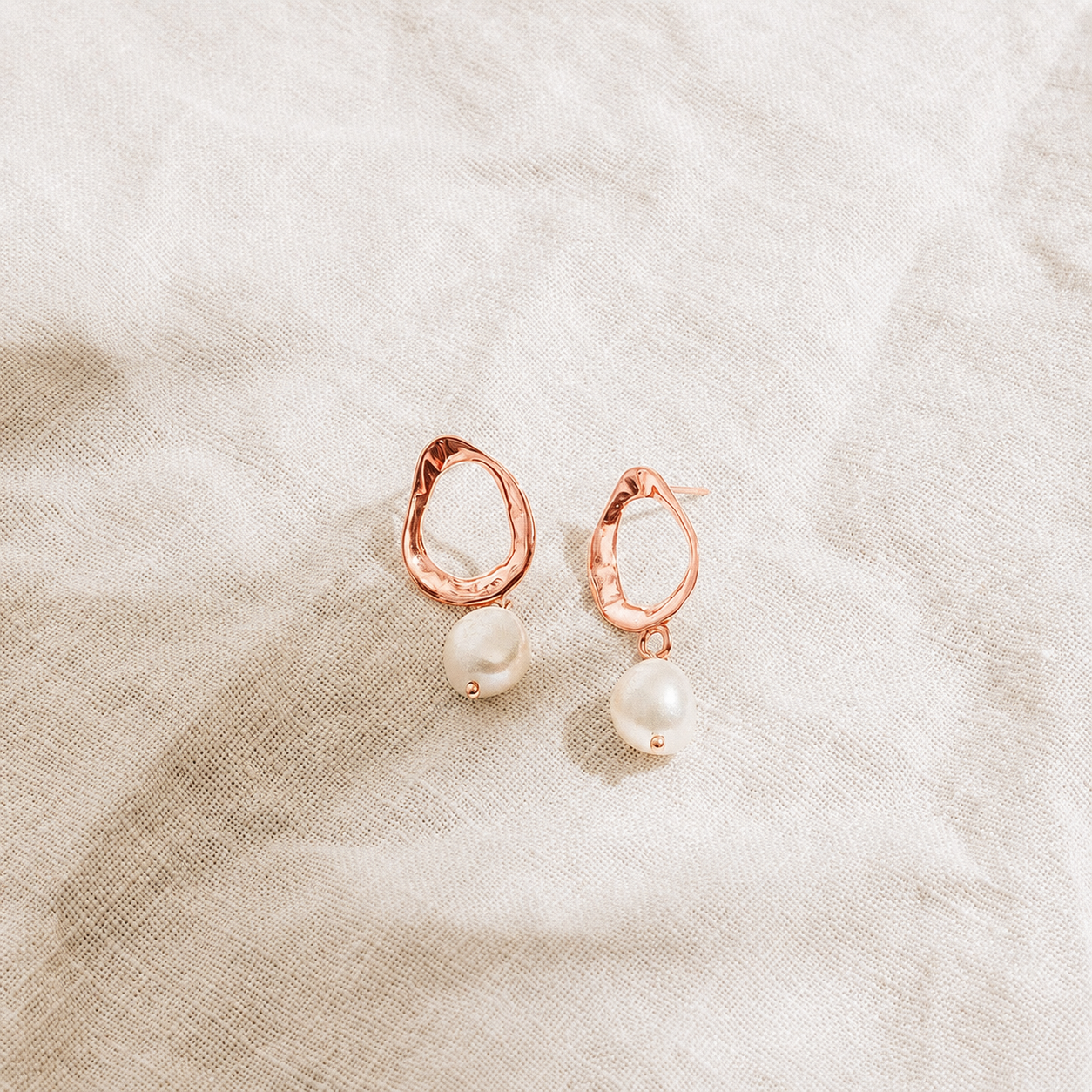 Rose gold earrings with pearl drops on a textured beige fabric background
