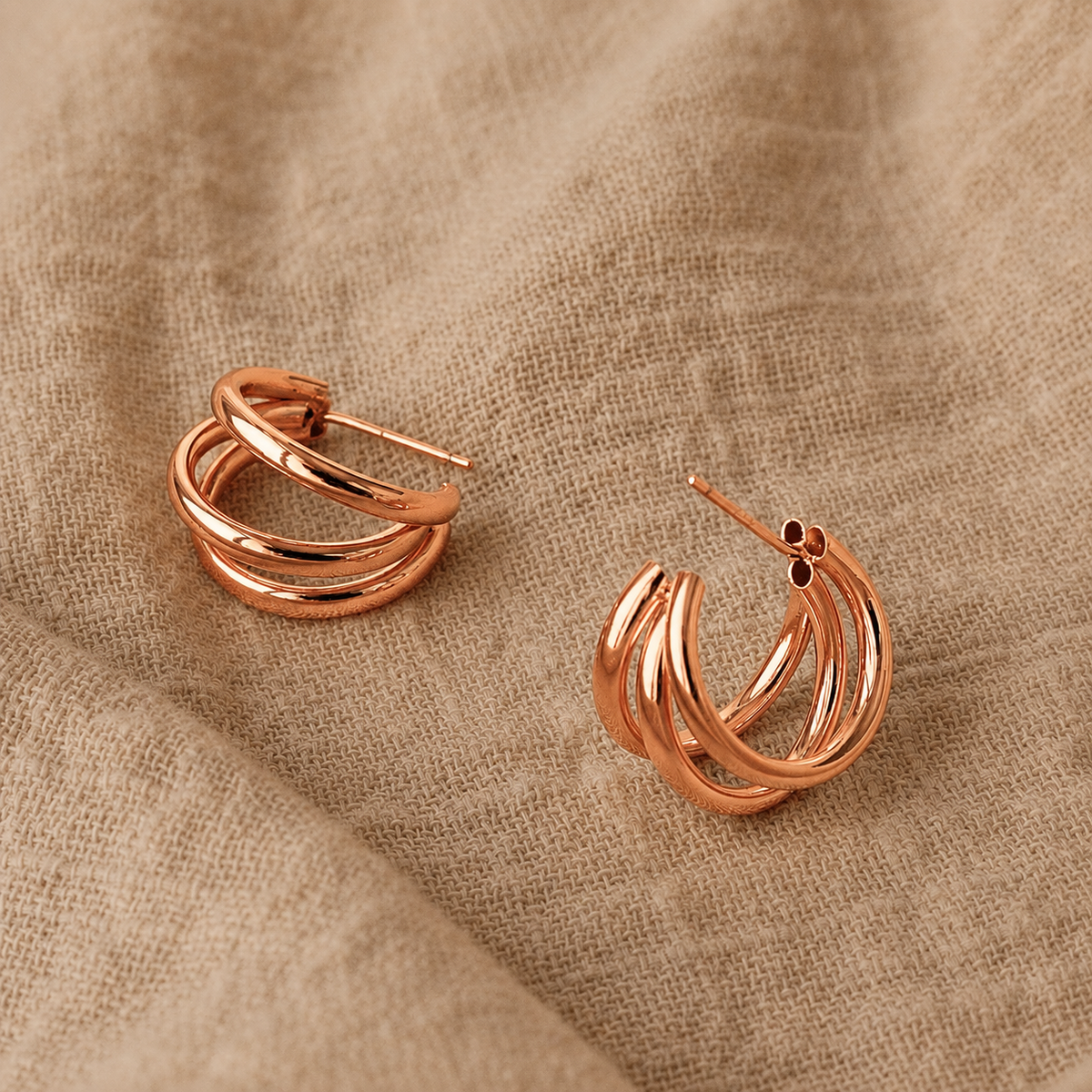Rose gold triple-band hoop earrings displayed on a textured beige linen fabric background.