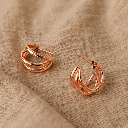 Rose gold triple-band hoop earrings displayed on a textured beige linen fabric background.