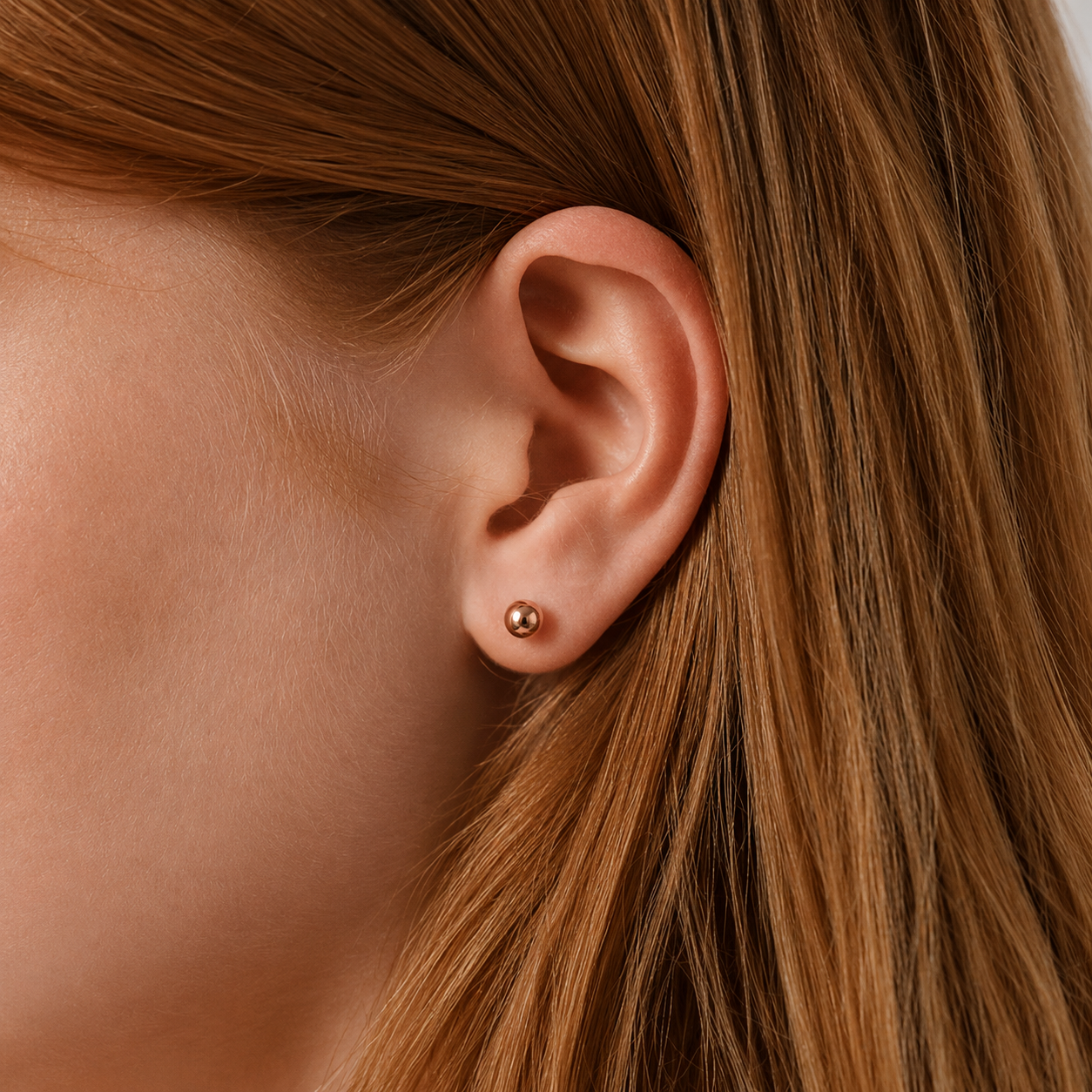 A close-up of a person's ear wearing a small round gold stud earring, with brown hair framing the side of the face.