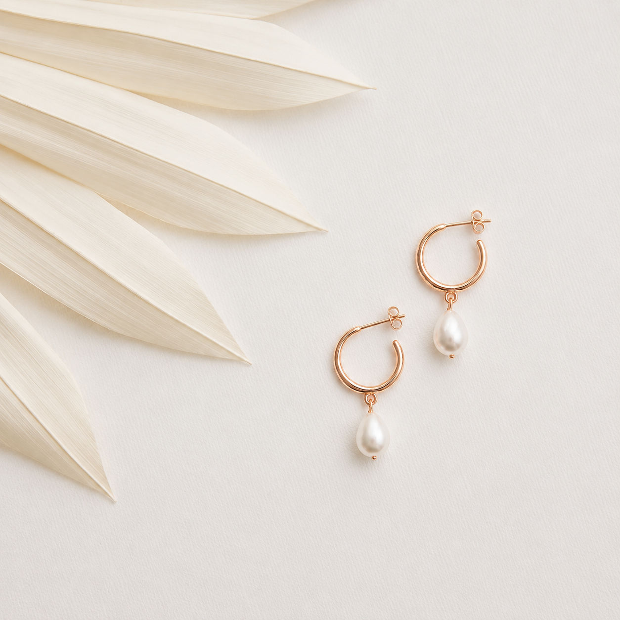 Rose gold hoop earrings with pearl drops on a light background with dried leaves.