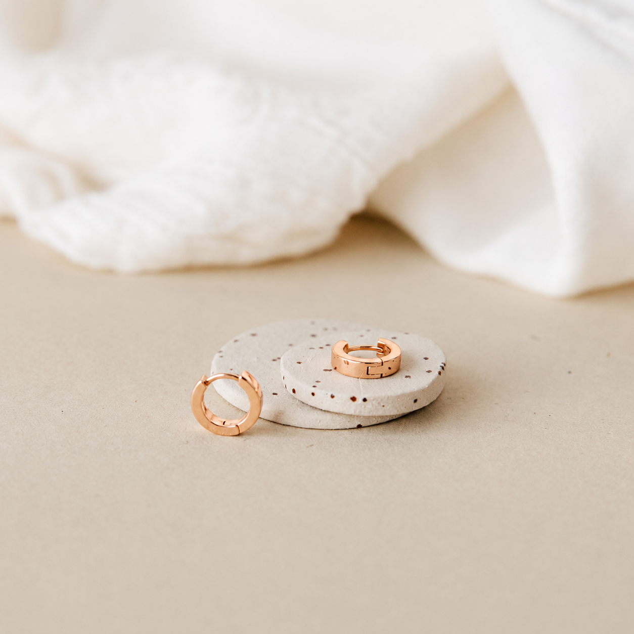 Two rose gold rings displayed on stacked speckled ceramic discs with a soft white fabric backdrop on a beige surface.