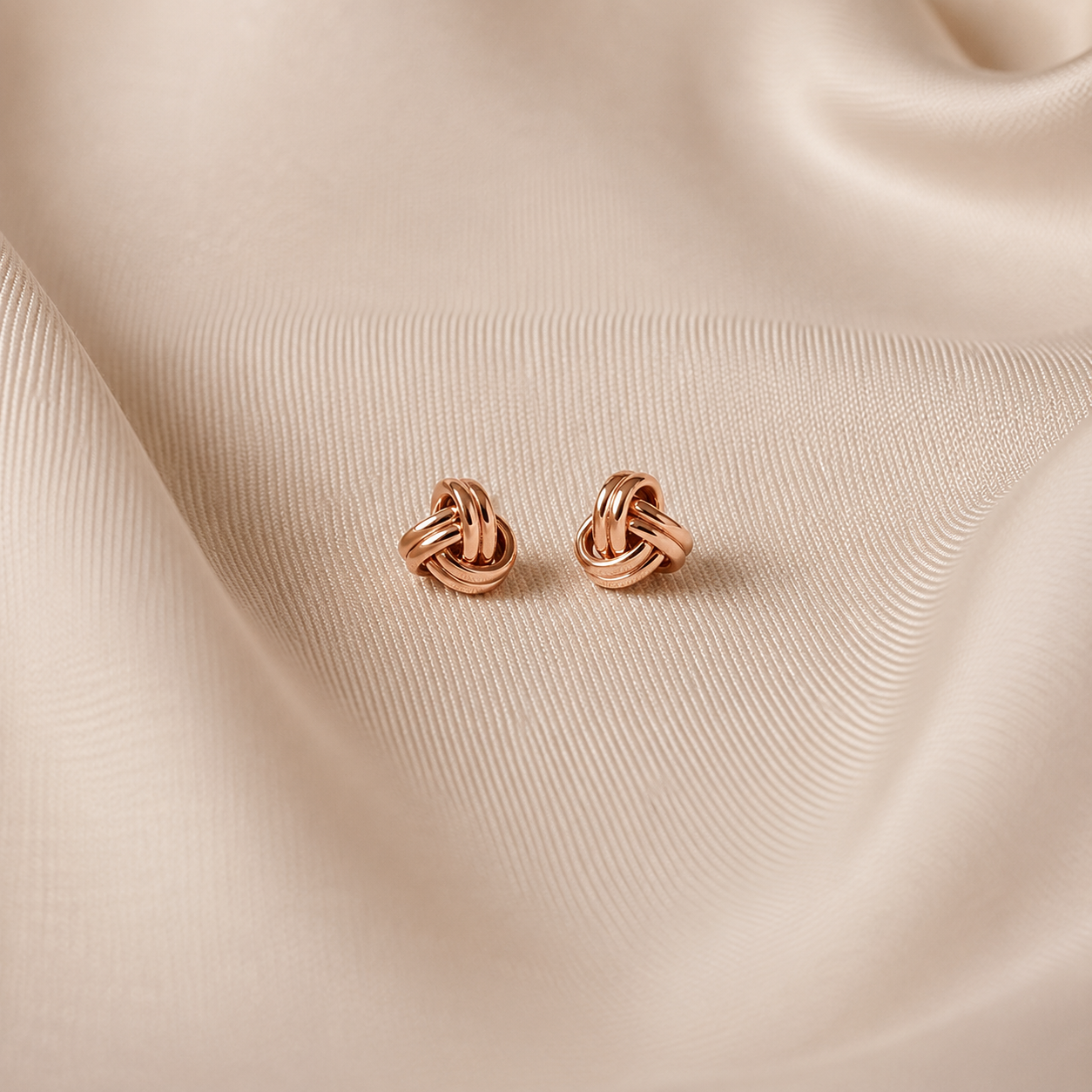 A pair of small rose gold knot stud earrings resting on draped beige satin fabric.
