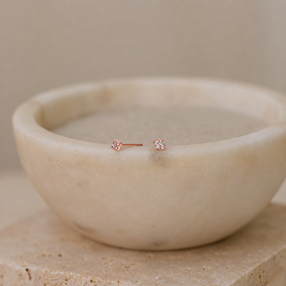 A pair of small rose gold stud earrings with clear gemstones, resting on the rim of a light beige stone bowl.