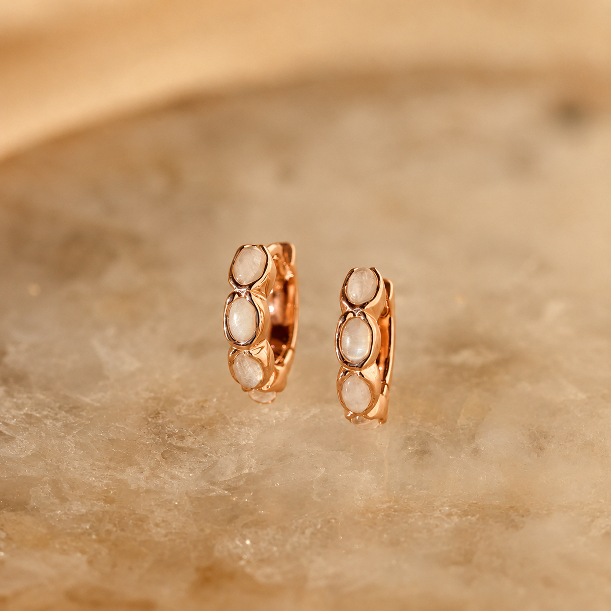 Rose gold hoop earrings with embedded stones on a beige stone background