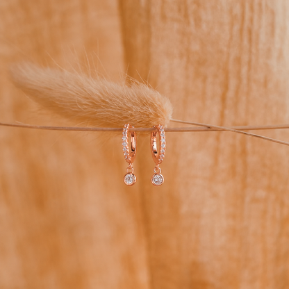 Rose gold hoop earrings with small round stones on a wooden surface with a feather.