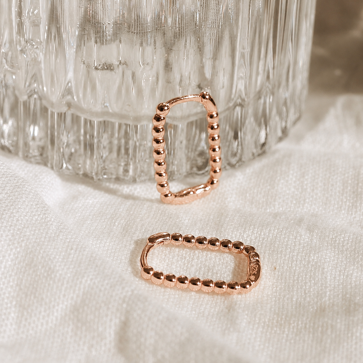 Rose gold hoop earrings on a textured white surface with a glass container in the background.
