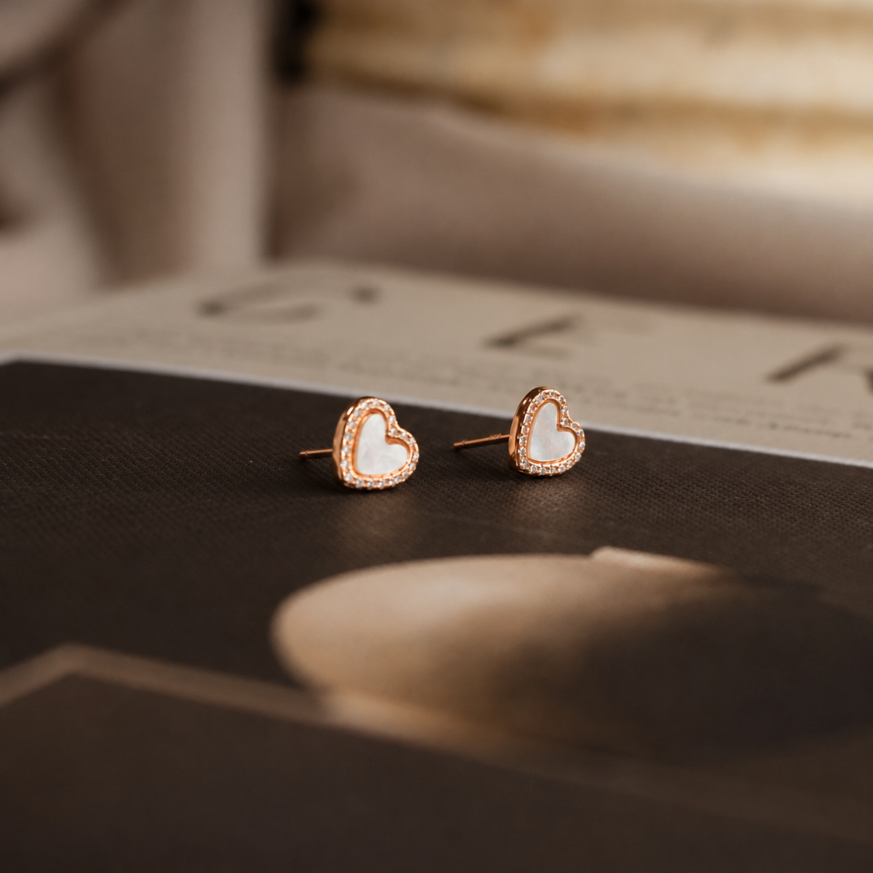 A pair of small heart-shaped stud earrings with white inlay and a rose gold border, resting on a dark textured surface with a blurred magazine in the background.