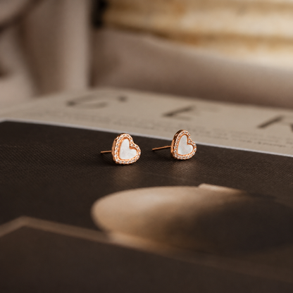 A pair of small heart-shaped stud earrings with white inlay and a rose gold border, resting on a dark textured surface with a blurred magazine in the background.