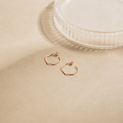 Rose gold hexagonal hoop earrings on a beige surface with a white dish in the background.