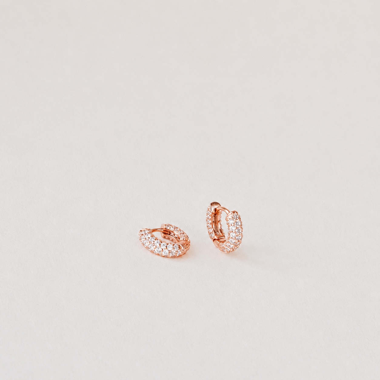 A pair of rose gold huggie hoop earrings paved with small clear stones, displayed on a clean, light beige background.
