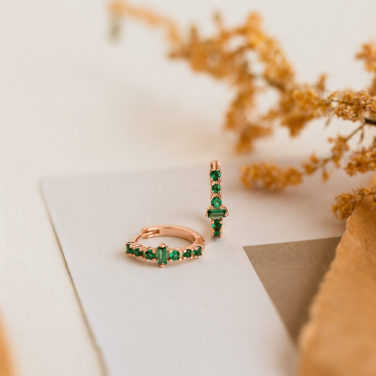 Rose gold rings with green gemstones on a white surface with dried flowers.