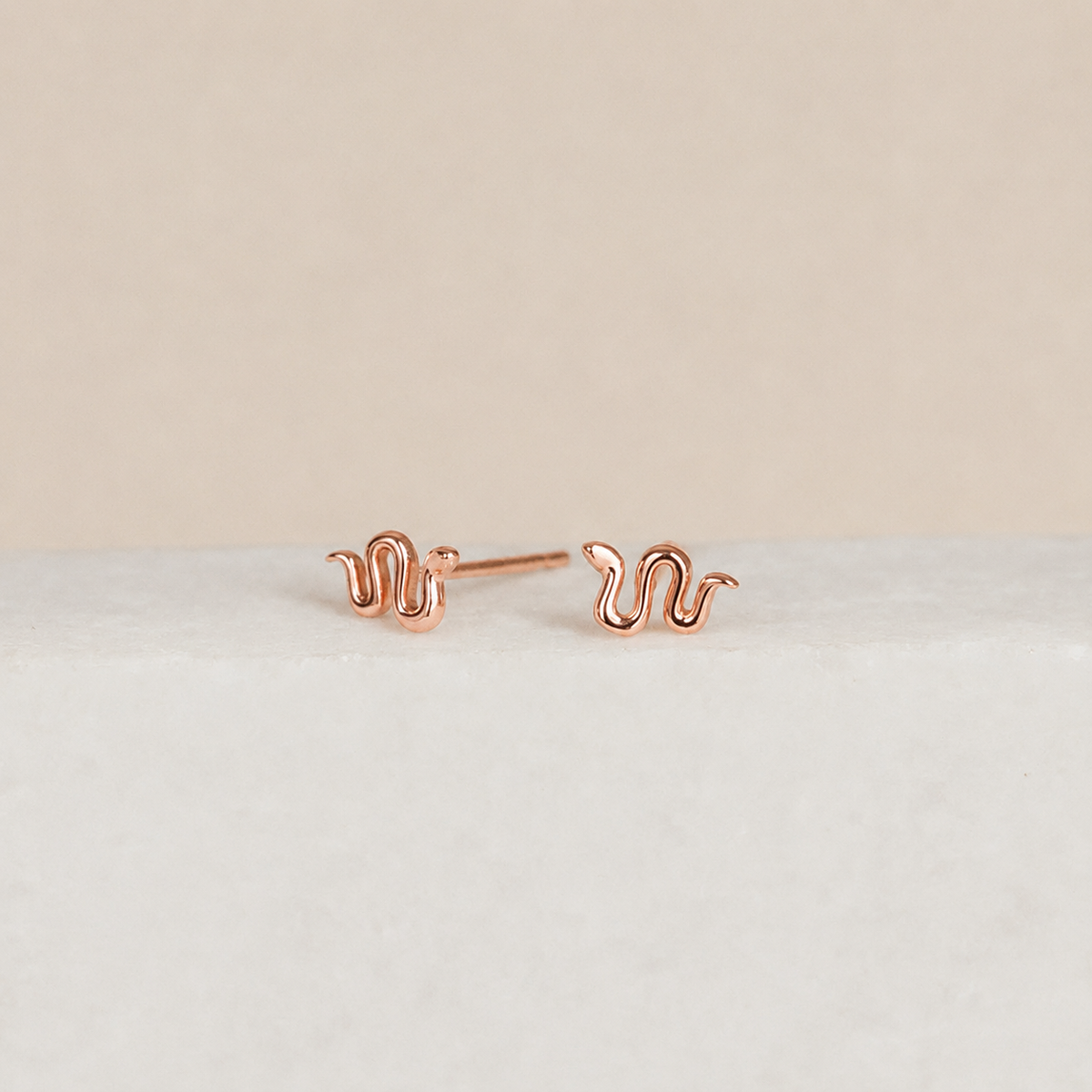 Rose gold snake-shaped earrings on a light beige background