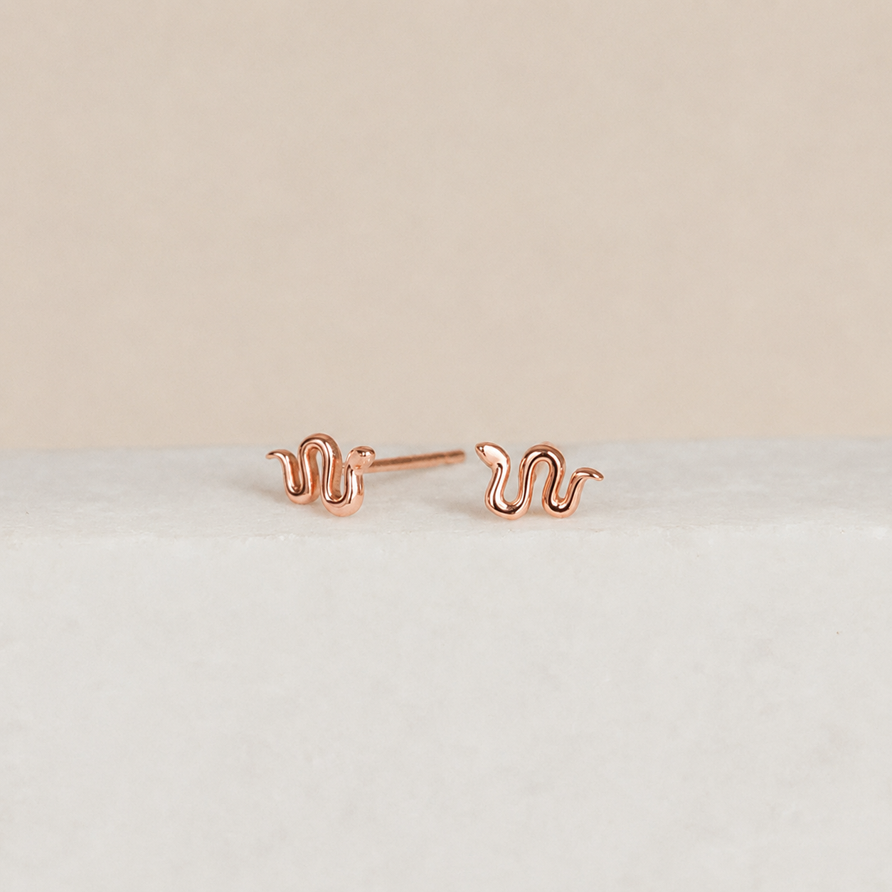 Rose gold snake-shaped earrings on a light beige background