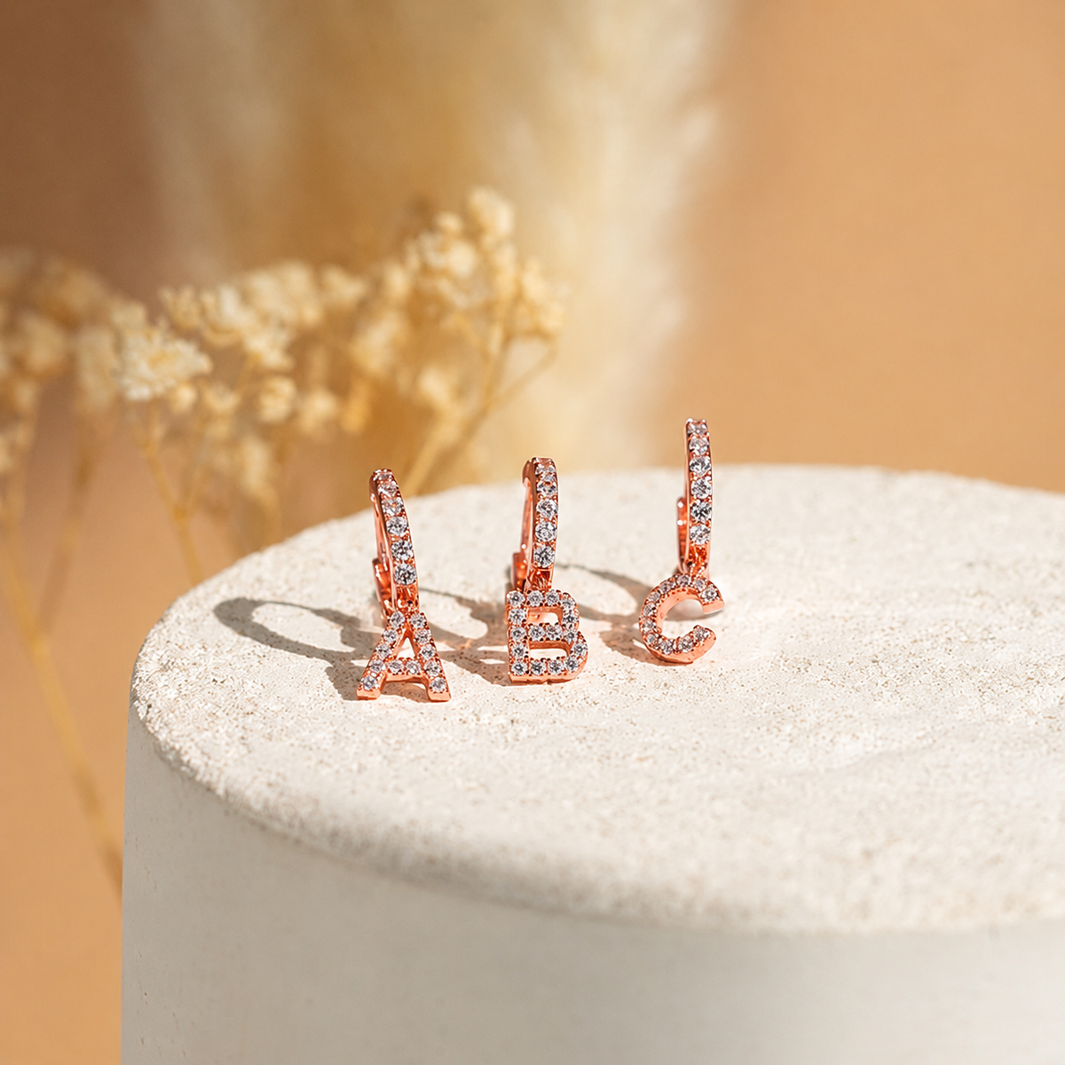 Rose gold letter 'A', 'B', and 'C' earrings on a textured surface with a blurred floral background.