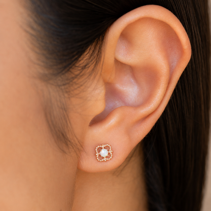 A close-up of a person's ear wearing a small square stud earring with a white pearl-like center and a rose gold floral halo.