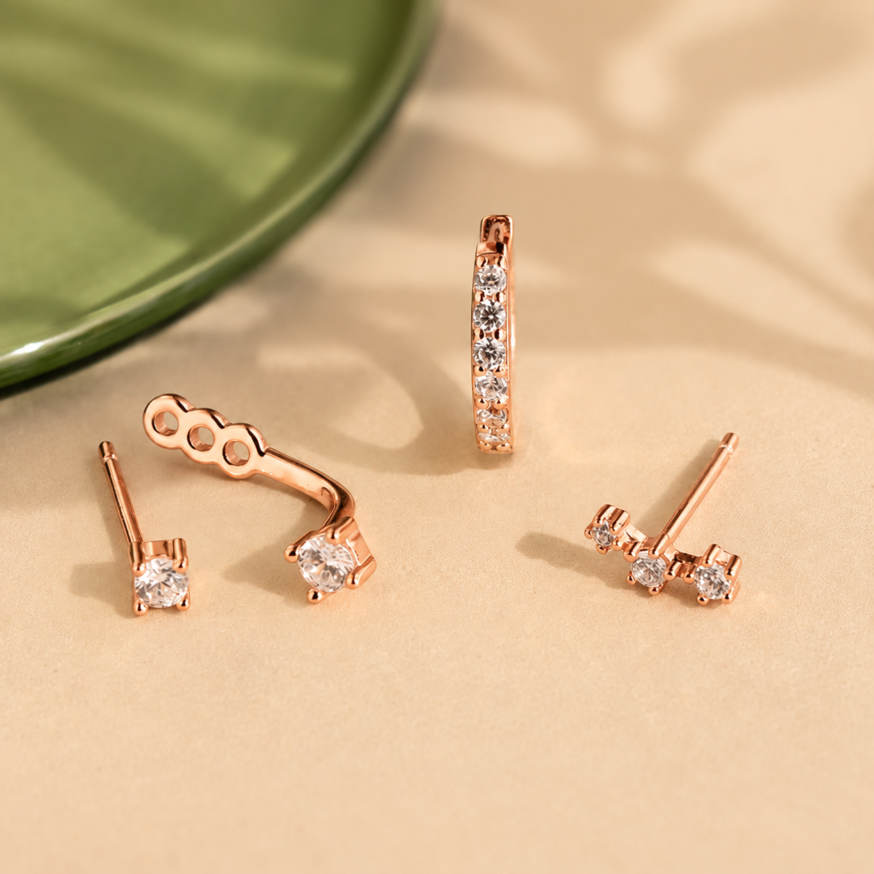 Set of rose gold earrings on a beige surface with a green plate in the background