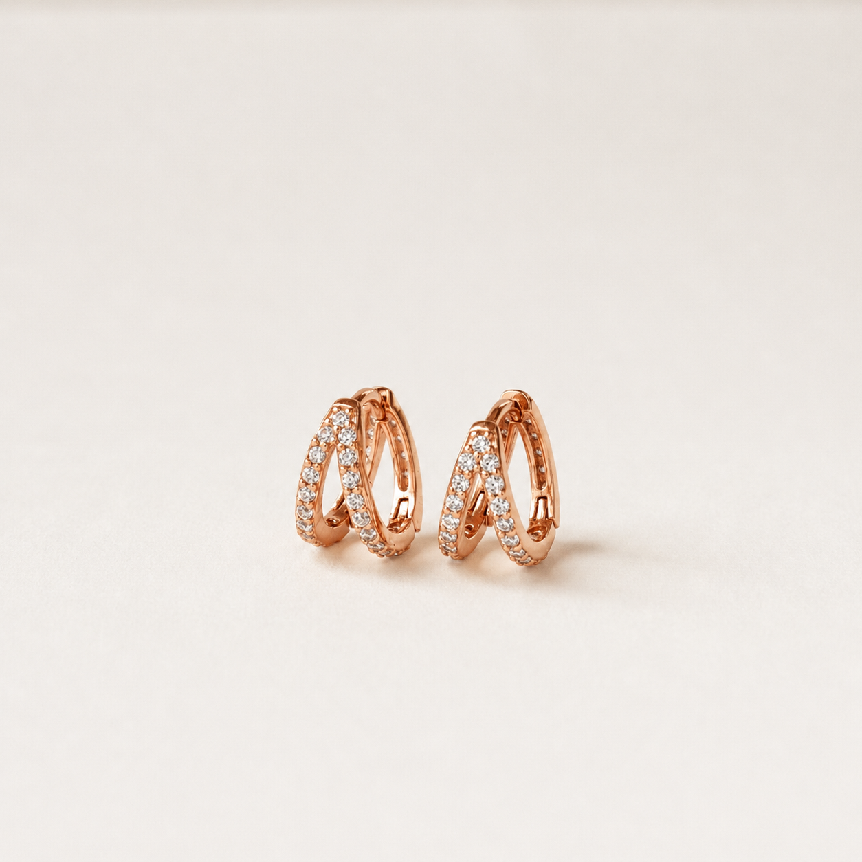 A pair of rose gold huggie hoop earrings with pavé-set clear stones, displayed on a clean, light beige background.
