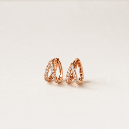 A pair of rose gold huggie hoop earrings with pavé-set clear stones, displayed on a clean, light beige background.