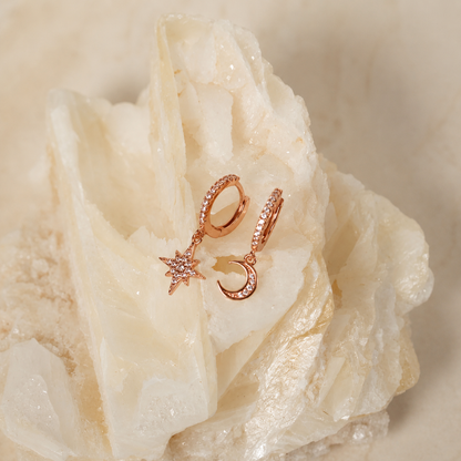 Rose gold crescent moon and star huggie hoop earrings with small clear crystal accents, displayed on a white crystal cluster.