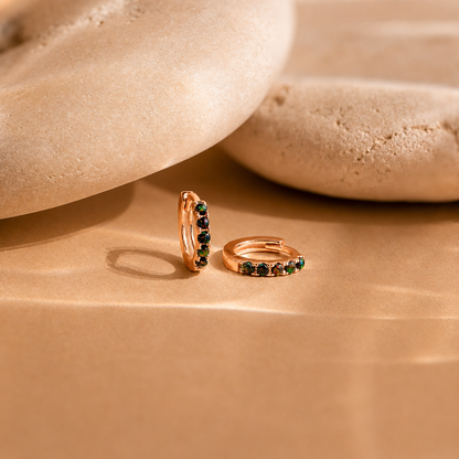 Rose gold hoop earrings with green gemstones on a beige stone surface