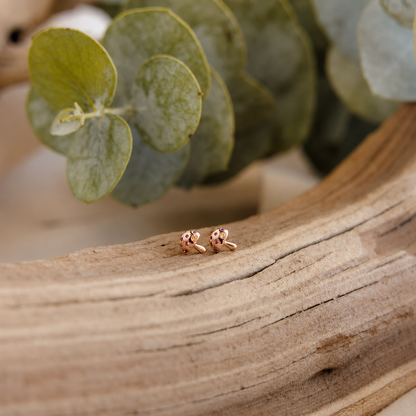 Rose gold bow-shaped earrings on a wooden surface with green leaves in the background