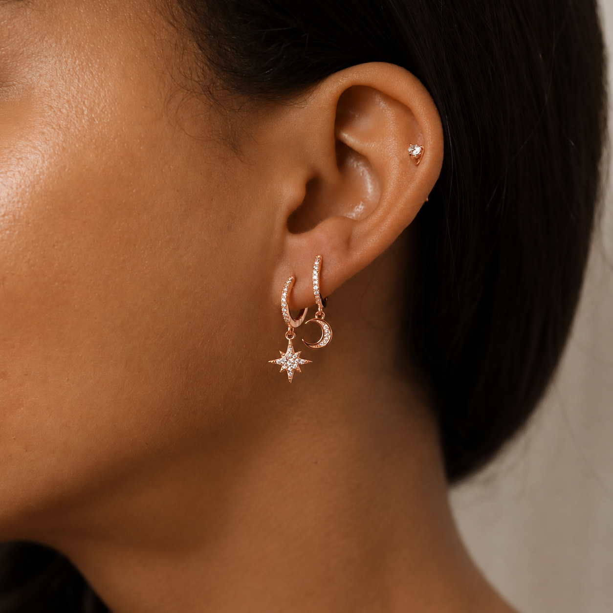 Close-up of a model's ear wearing a rose gold huggie hoop earring with a dangling star and crescent moon charm, alongside a small stud earring.